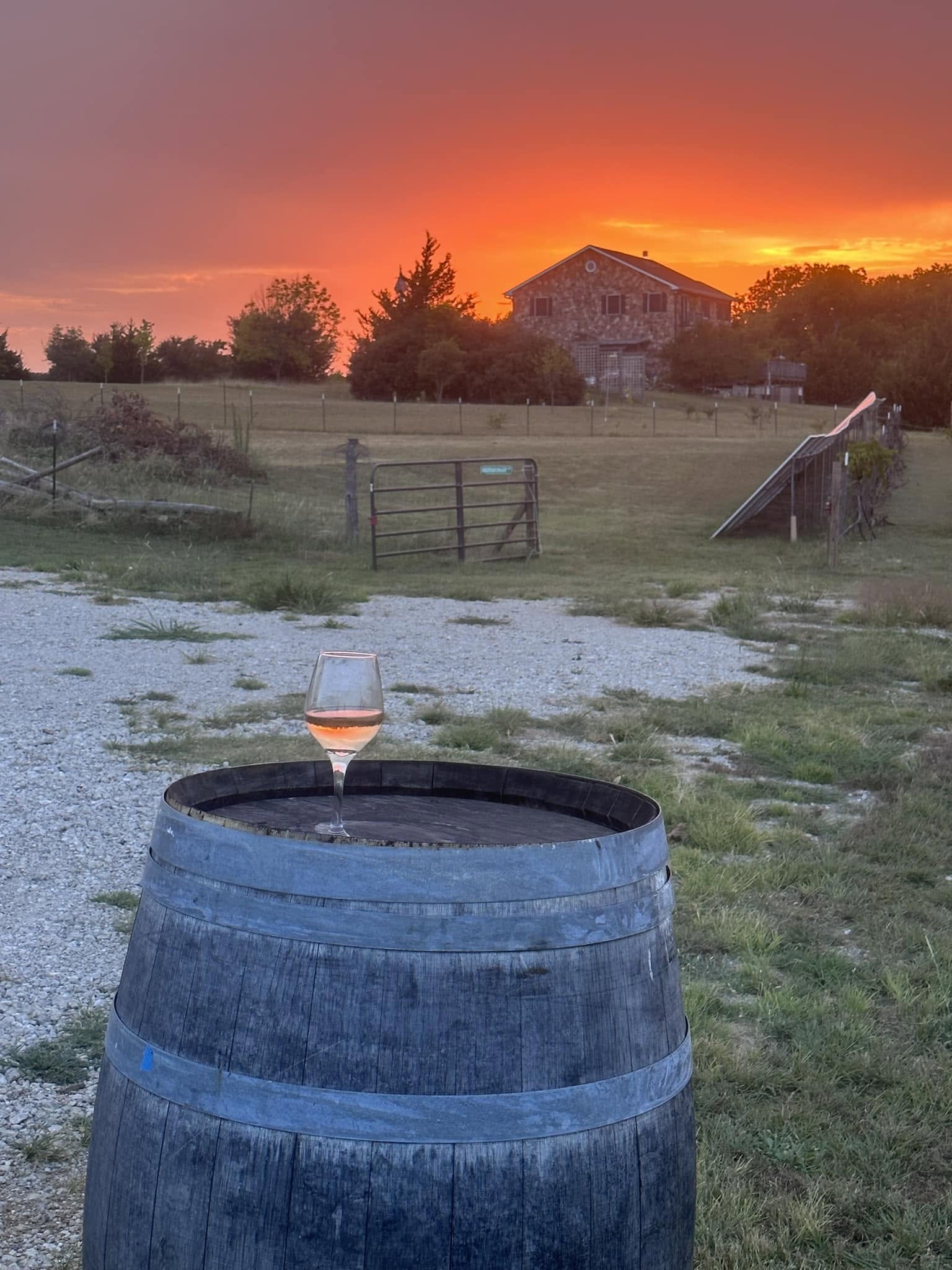 A glass of wine on a barrel with the rustic barn and sunset behind it